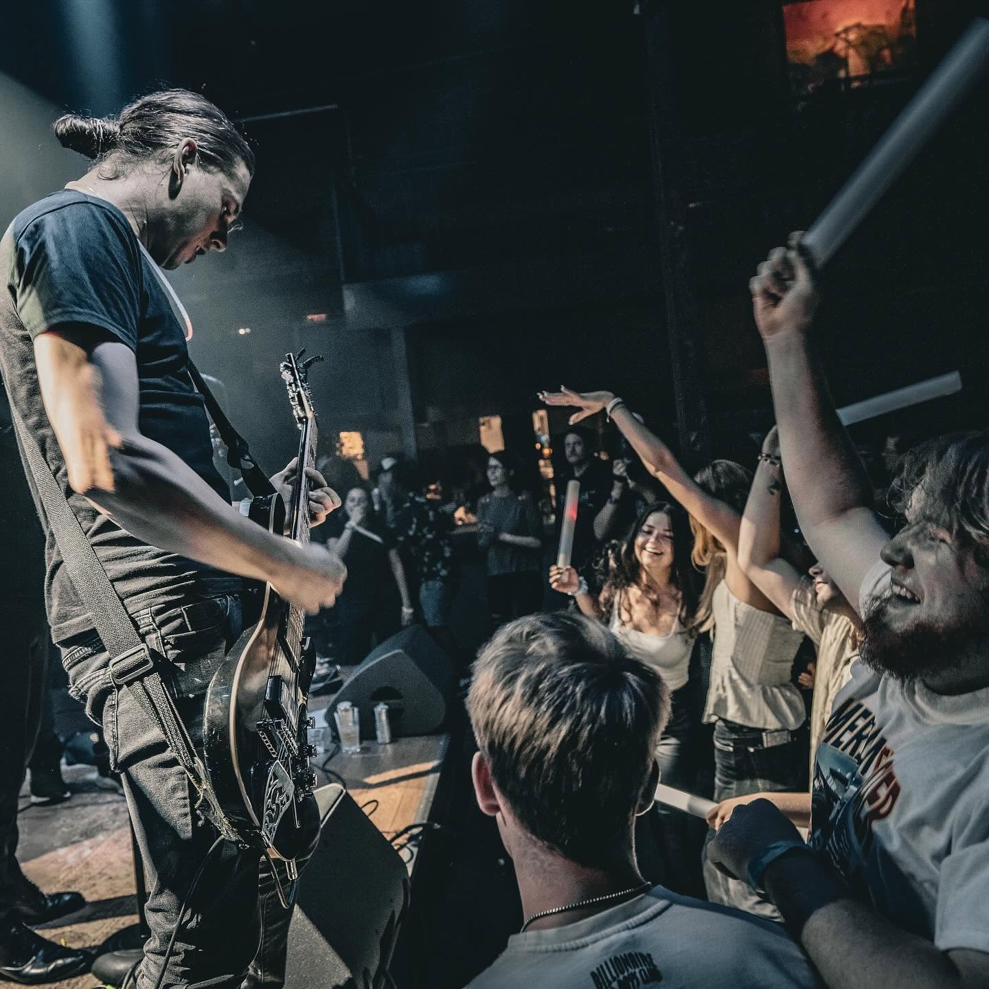 Musician playing guitar on stage with audience cheering and raising hands in a dark concert venue.